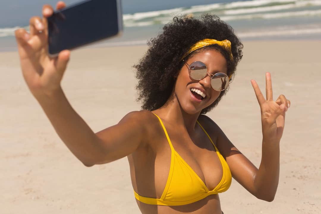 Woman on a beach in a bikini with ideal body that can be attained via cosmetic surgery, making a peace sign and taking a selfie.