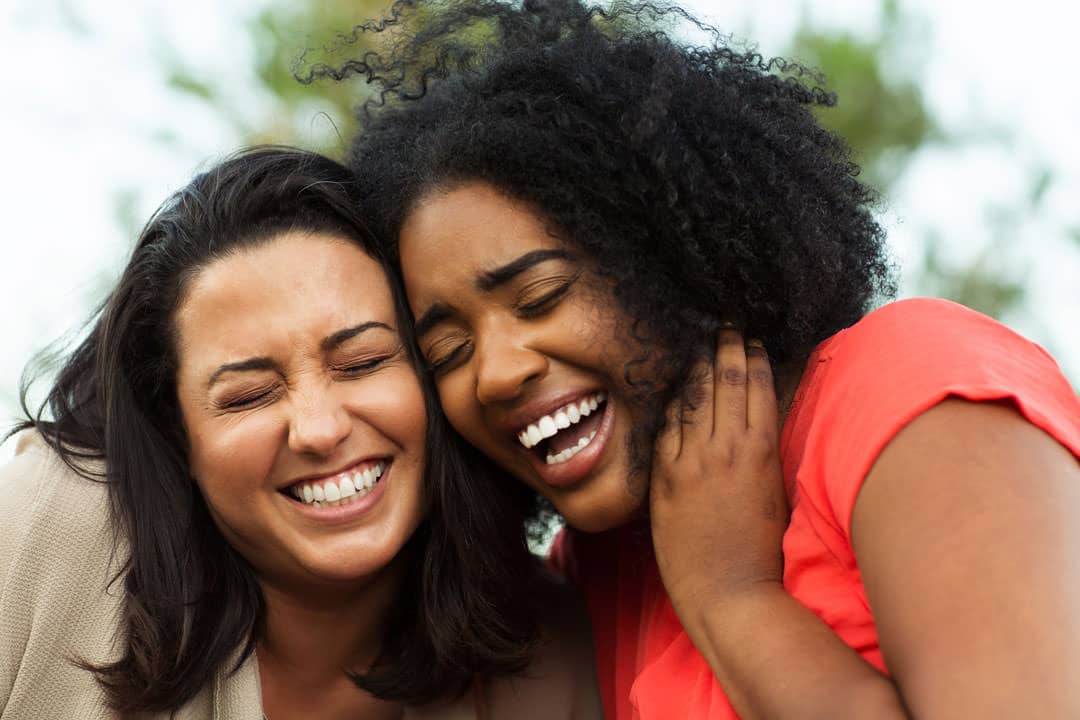 Headshots of two smiling women with heads together and eyes closed, appear to be laughing.